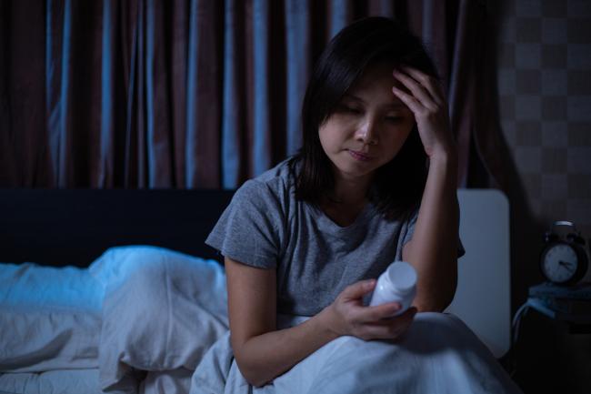 20220301-melatonin.jpg Woman sitting up in bed and looking at a pill bottle.
